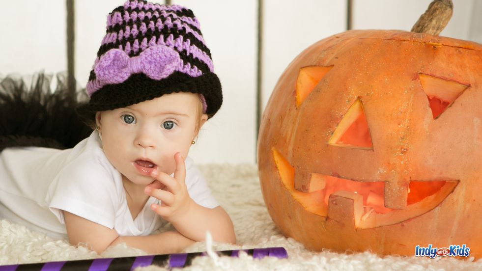 A baby in a hat poses with a carved jack-o-lantern pumpkin.