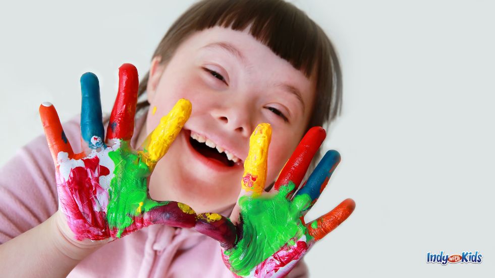 A child with Downs Syndrome smiles while holding up her colorful, paint-covered hands.