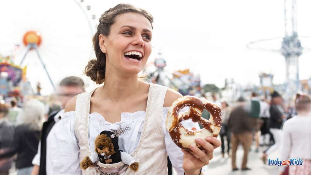 Indianapolis Events OCTOBER EDITION: a woman in German gear at an Oktoberfest event holds a Bavarian pretzel with a festival blurred behind her