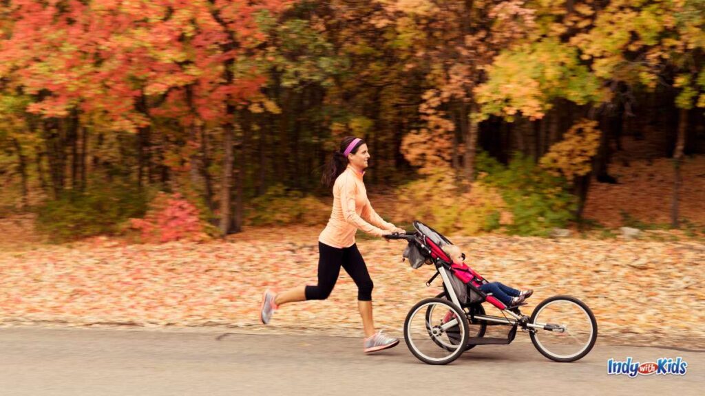 20 Great Things to Do in Indianapolis in November: a woman jogs and pushes a red stroller with a toddler inside on a trail with a leaf covered ground and orange/green/yellow leaves behind her