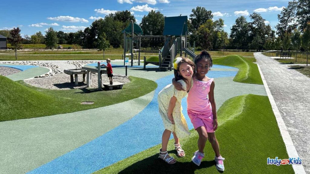 two little girls hug in front of the playground at Virgil Park. there is a boy at a water table in the background.