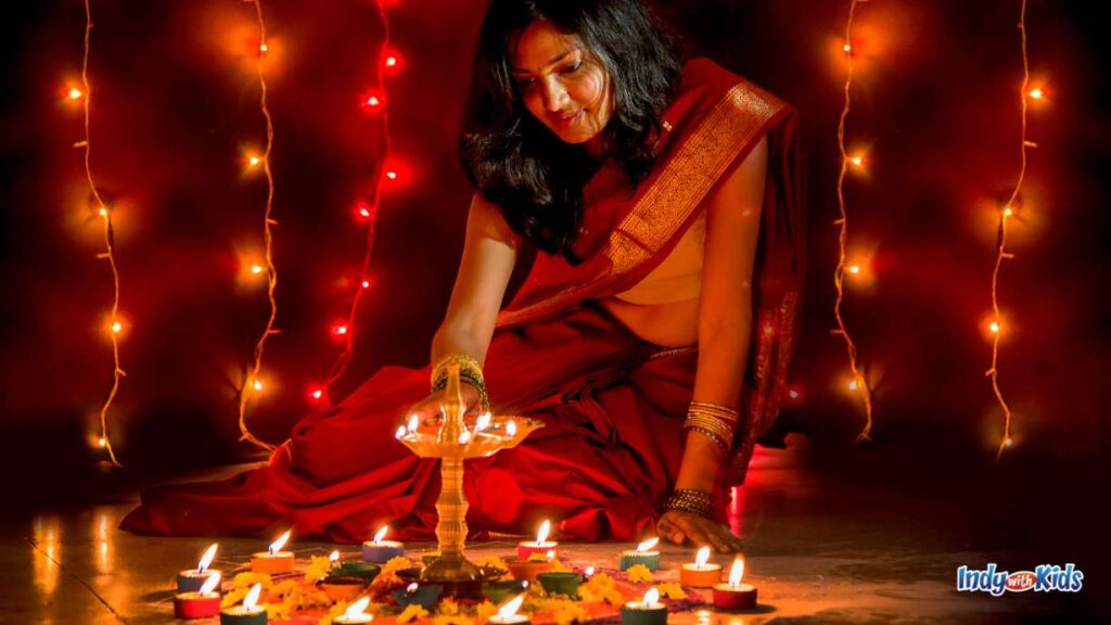 a woman in a red sari sits on the floor in front of several tea light candles and lights a candle atop a tall plate for diwali