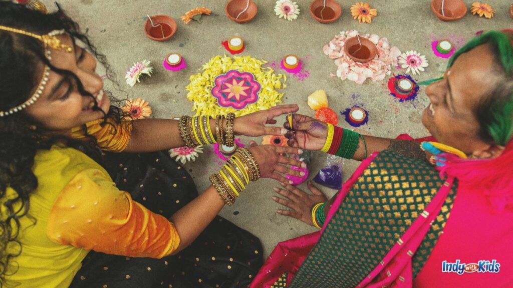 a woman and a older woman smile at each other, each dressed in saris and sitting amonst rangolis made of sand and tea light candles for diwali