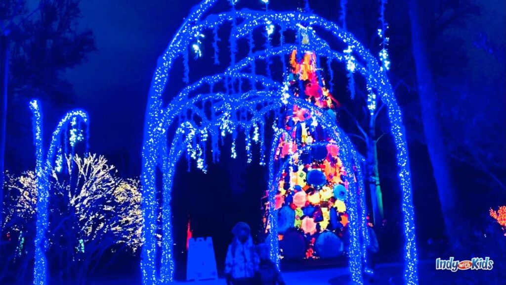 arched branches over two children are covered in lights at Winterlights at Newfields. there is a colorful tree in the background lit up.