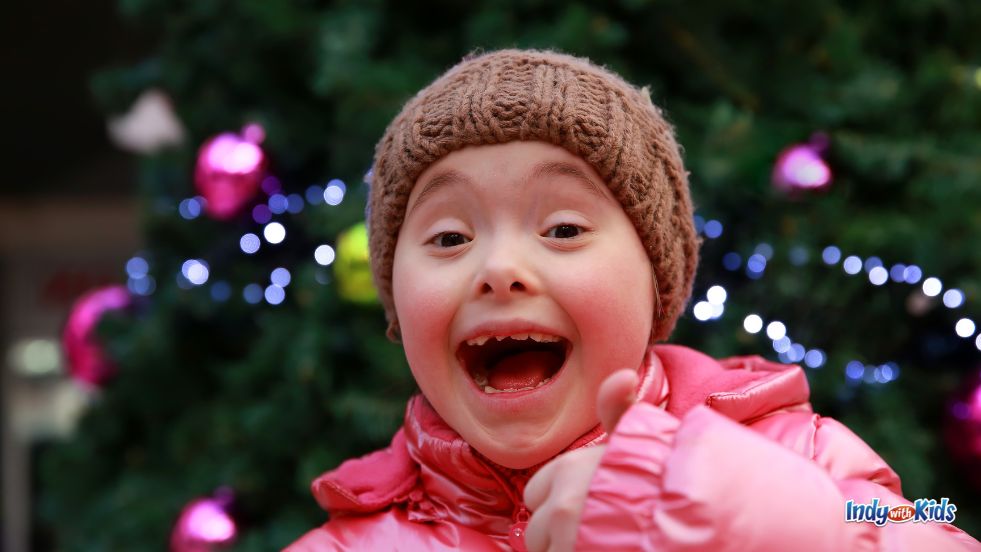 A girl wearing a pink coat and a brown hat grins and gives a thumbs up to the camera while posing in front of Christmas lights. 