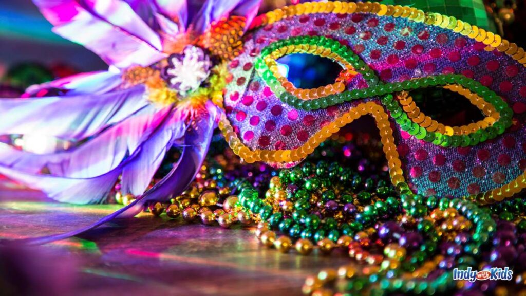 a masquerade mask featuring lots of purple green and yellow with feathers coming out of one side sits on a table behind a handful of bead necklaces