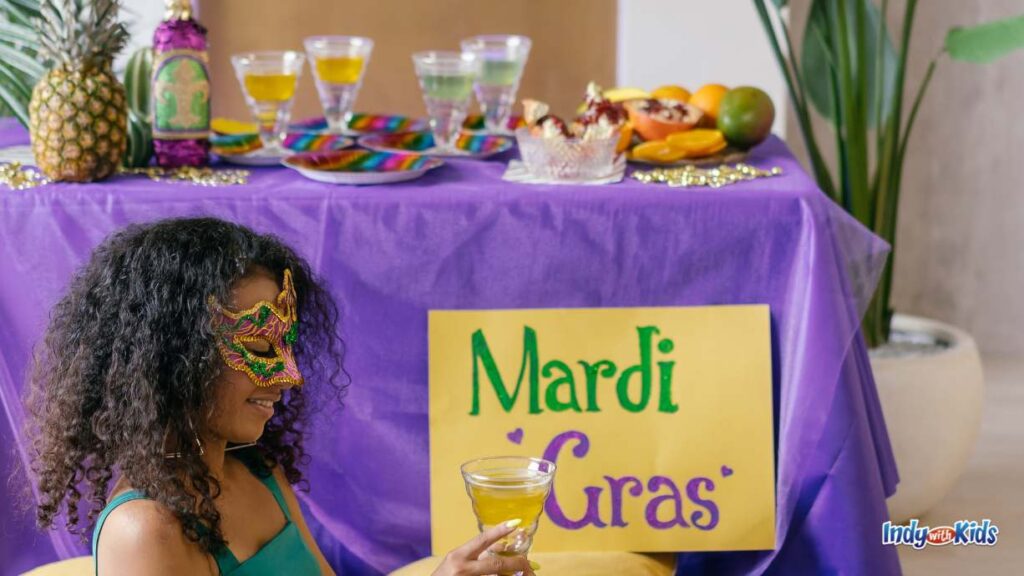 a woman with a maquerade mask sits on the floor with a drink in front of a table that has a Mardi gras sign. there are Mardi gras inspired food and drink on the table.