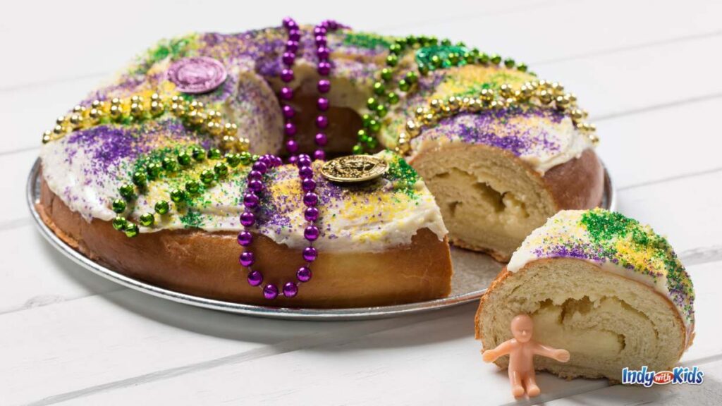 A Mardi Gras king cake with icing, purple green and yellow beads, and coins on top. a slice has been taken out and a tiny baby figurine is sat up against it.