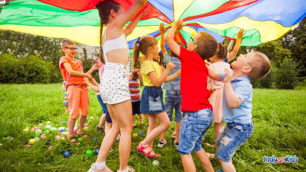 Spring Break Camp Indianapolis: several children play under a primary color parachute as it floats in the air