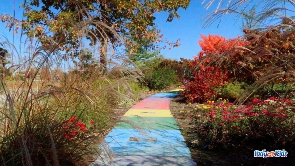 a path of rainbow colored tiles leads the way through a garden of flowers bushes and plants on either side of it at coxhall gardens