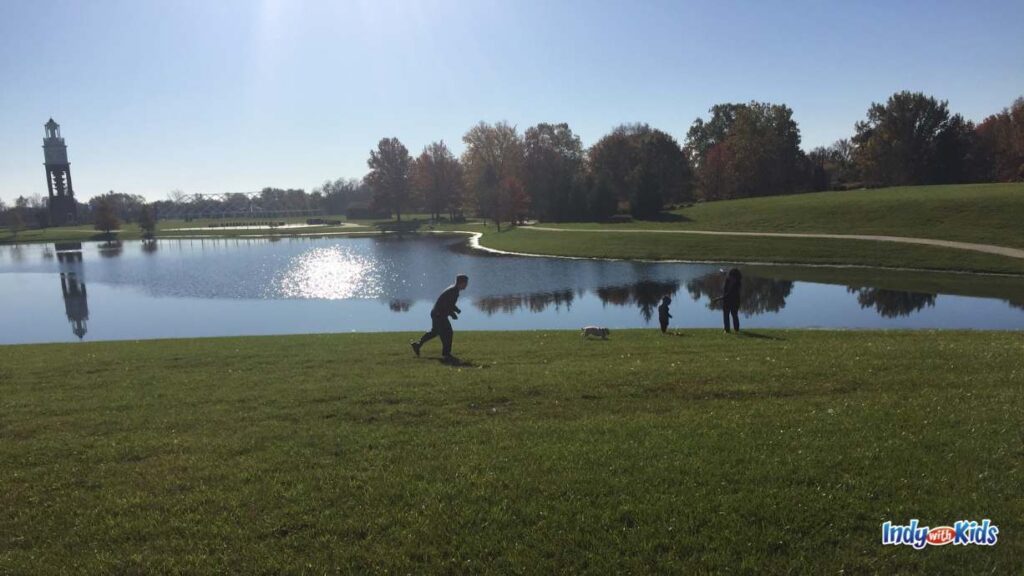 a bell tower sits in the distance being a large pond with walking trails around it and a grassy field in the foreground