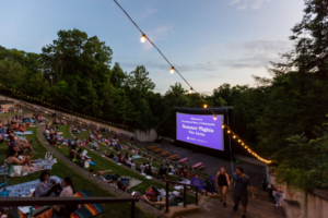 Families spread blankets across the grassy amphitheater at Newfields in Indianapolis for the Summer Nights Film Series, with string lights glowing above the outdoor movie screen at dusk.