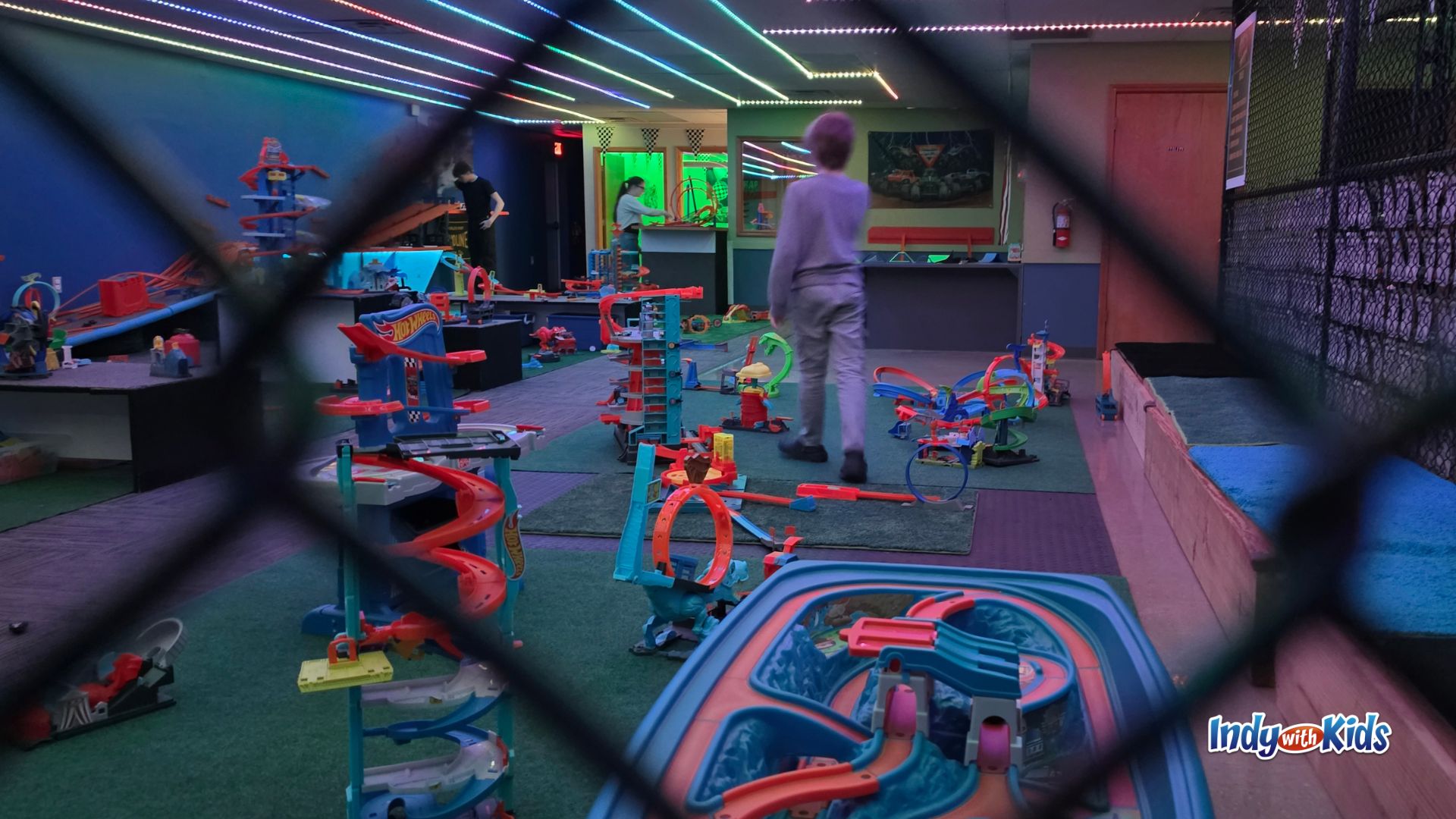 View through safety fencing into SpeedZone play area with toy race tracks and a child walking through the room.