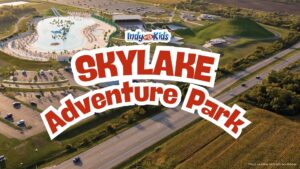 Conceptual aerial rendering of SKYLAKE Adventure Park showing a large swimming lake with sandy beaches, water attractions, buildings, parking areas, and grassy hills. Text overlay says “SKYLAKE Adventure Park.”