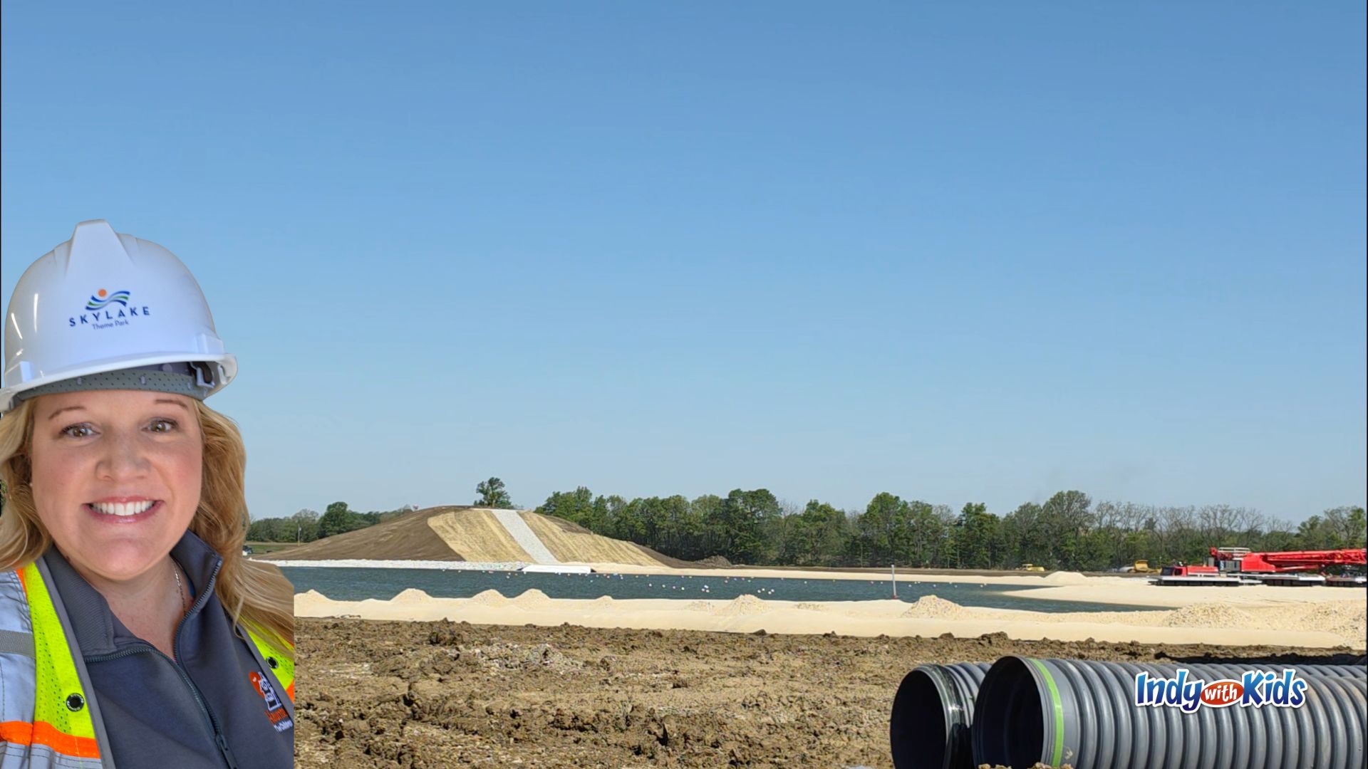 Katy Mann wearing a SKYLAKE hard hat and safety vest during a construction site preview, with the new lake, white sand shoreline, and large slide hill visible behind her.