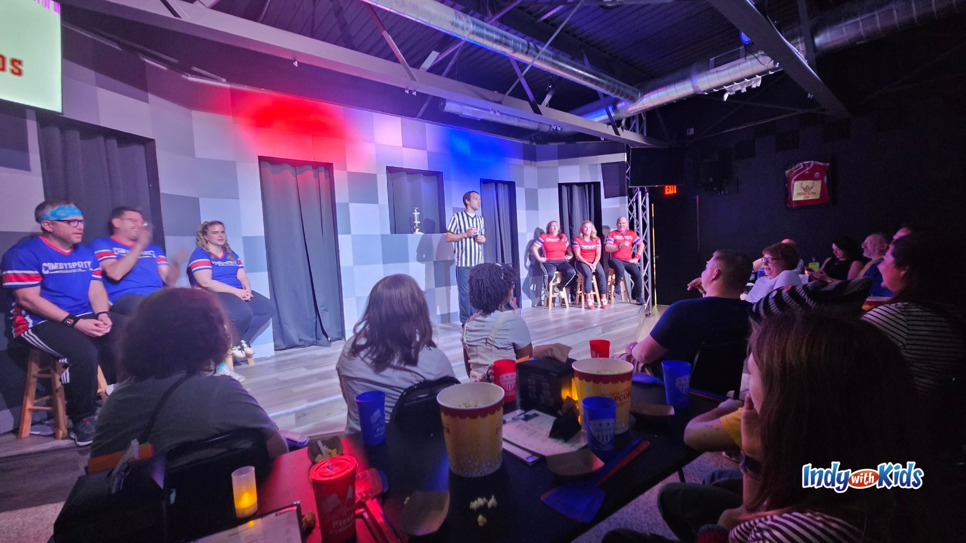 Audience members watch an improv comedy performance at ComedySportz Indianapolis, where performers in red and blue team shirts sit onstage beside a referee under colorful theater lights.