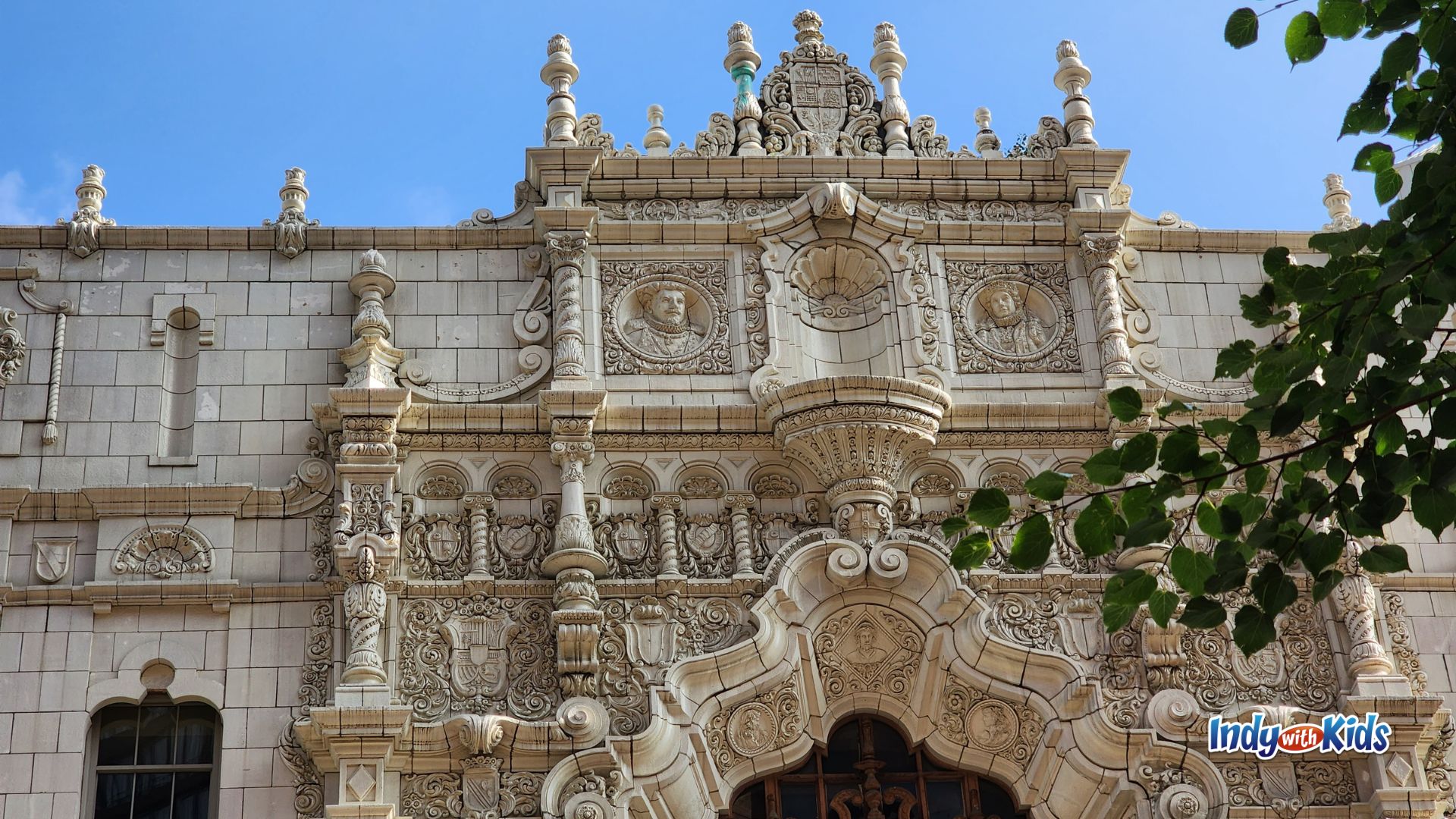 Close-up of an ornate historic building facade in downtown Indianapolis, (The Indianapolis Repertory Theater) covered with detailed stone carvings, decorative columns, scrollwork, and sculpted faces beneath a bright blue sky.