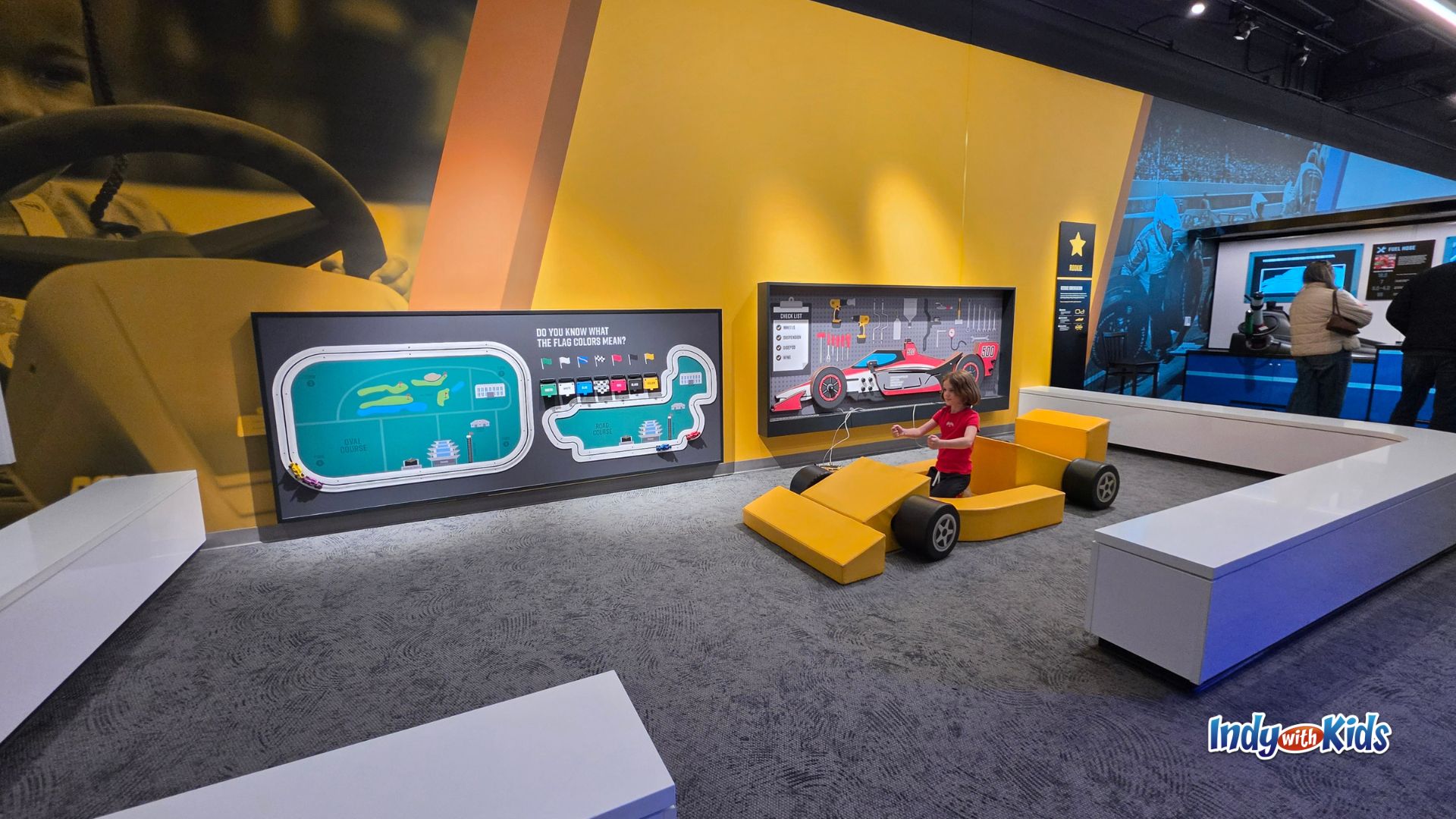 A child plays in a kid-sized yellow race car in the children’s play area at the Indianapolis Motor Speedway Museum, surrounded by interactive displays about race tracks, flag colors, and race car design.
