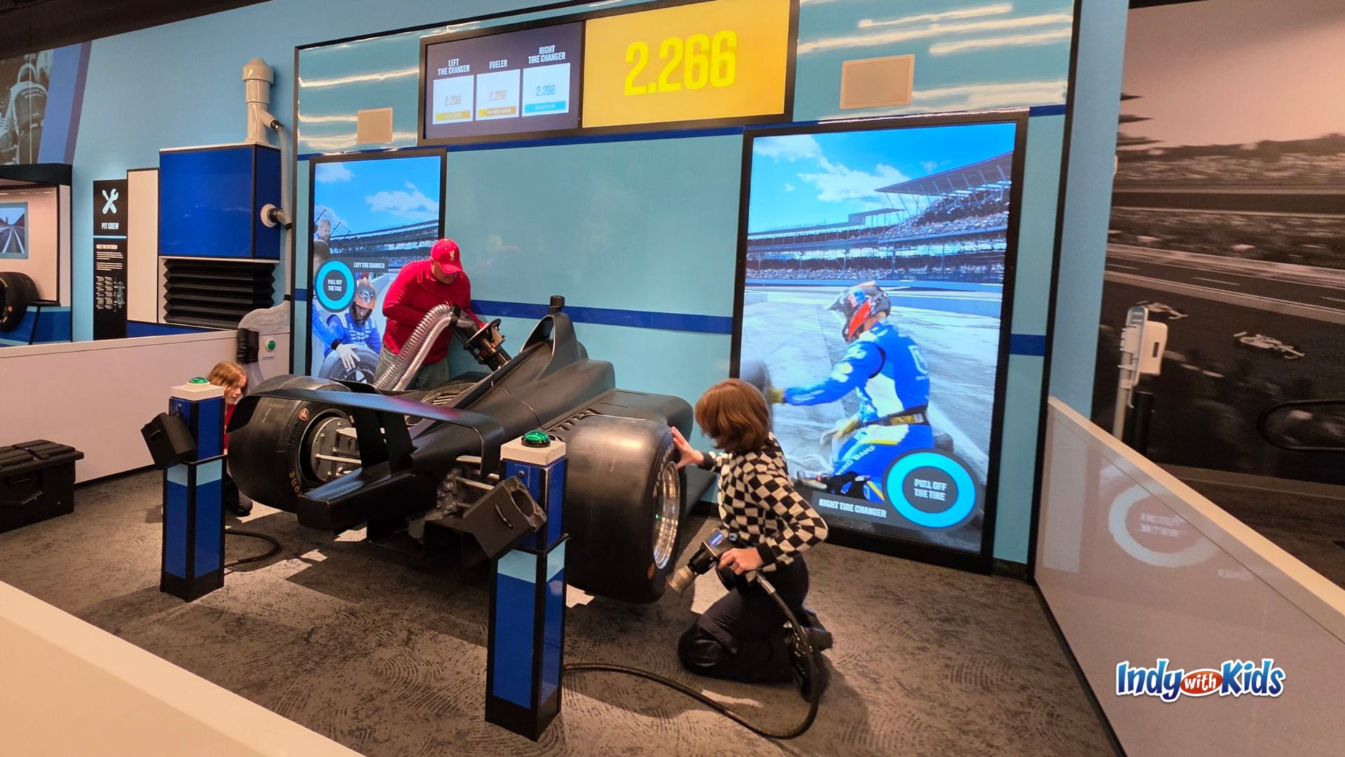 Two children and their father work together during an interactive pit stop challenge at the Indianapolis Motor Speedway Museum, changing tires and fueling a black Indy car while digital screens track their time.