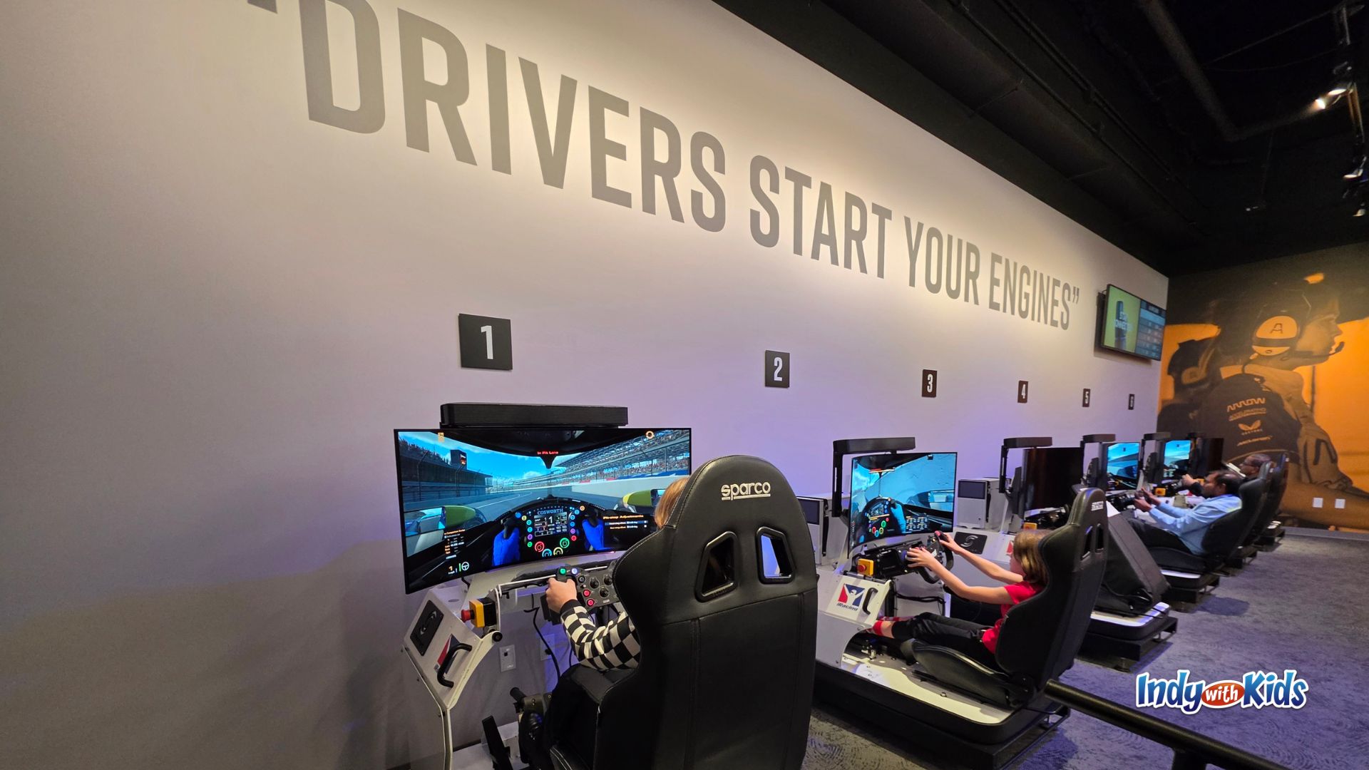 Kids race on driving simulators at the Indianapolis Motor Speedway Museum beneath large wall text that says, “Drivers Start Your Engines.”