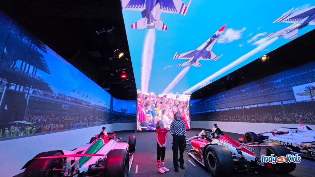 Two children stand among Indy race cars in an immersive gallery at the Indianapolis Motor Speedway Museum while giant screens show a flyover above cheering race fans at the track.