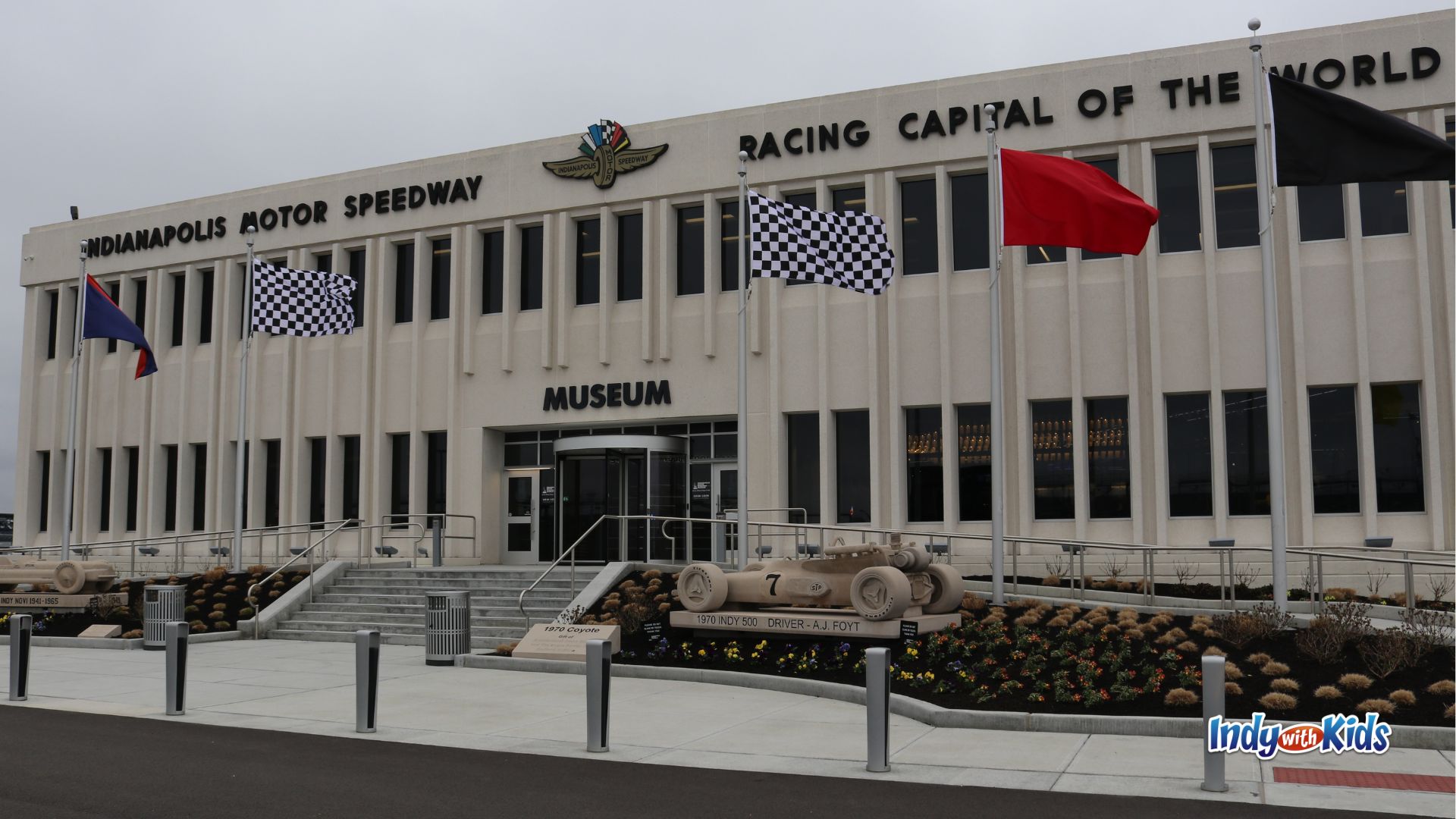 Front entrance of the Indianapolis Motor Speedway Museum with checkered flags, red and black racing flags, and a sculpture honoring A.J. Foyt’s 1970 Indy 500 race car.