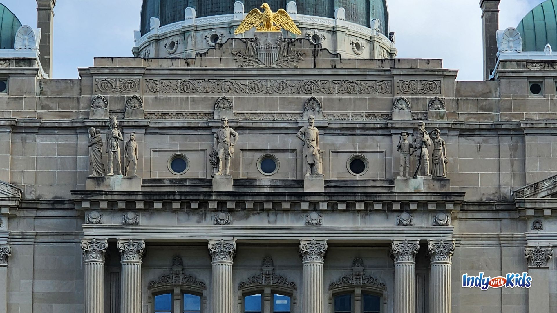 Upper facade of the Indiana State House in Indianapolis, specifically highlighting the limestone figures representing the "Westward Journey" created in 1887. featuring stone statues, classical columns, carved details, and a gold eagle above the entrance.