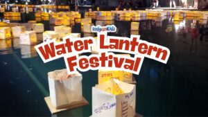 Hundreds of glowing paper lanterns float on the water at the Indianapolis Water Lantern Festival after dark, with visitors gathered along the edge of the pond. Text overlay reads “Water Lantern Festival.”