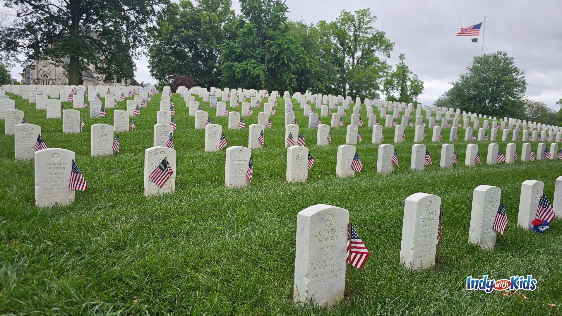 Rows of white military headstones decorated with American flags at Crown Hill National Cemetery in Indianapolis for Memorial Day, with a large American flag flying in the background.