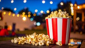 Red and white striped bucket overflowing with popcorn on an outdoor table at dusk, with string lights glowing in the background for a family-friendly outdoor movie night.