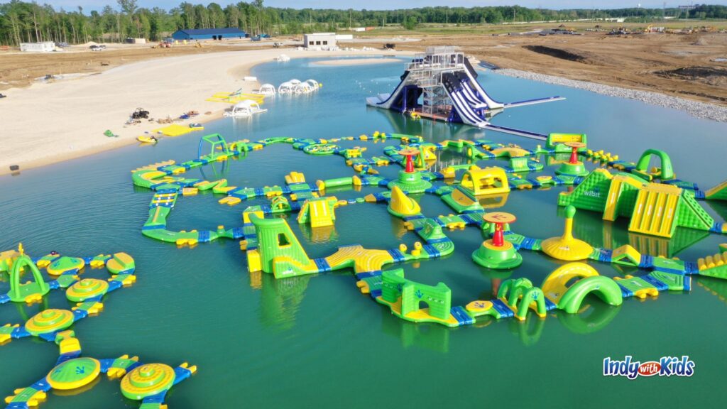 A large inflatable aqua park with green and yellow obstacle course features, slides, climbing structures, and floating play areas spread across a lake.