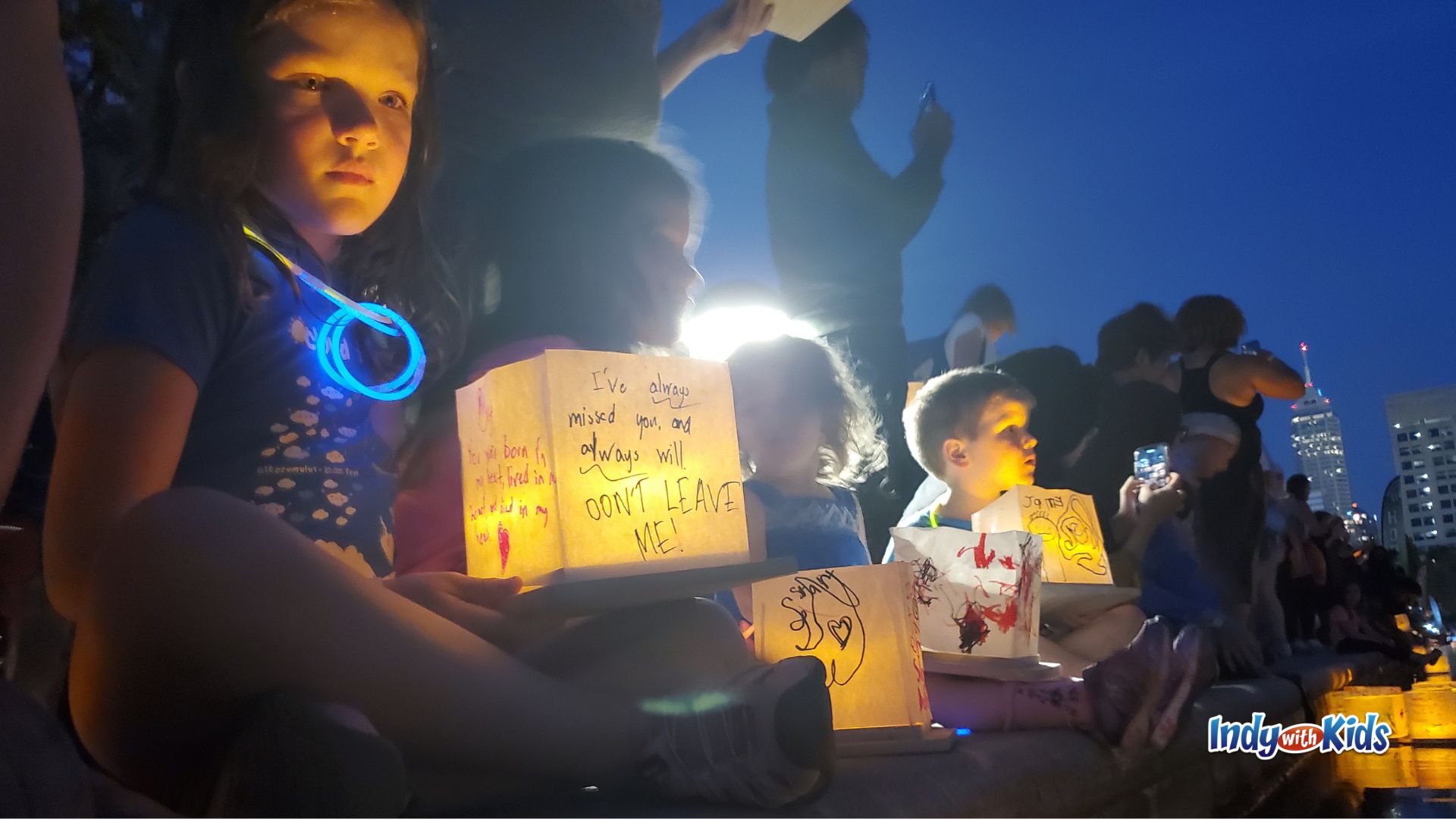 Children sit beside the water at dusk holding illuminated paper lanterns decorated with handwritten messages and drawings before the lantern release at the Indianapolis Water Lantern Festival.