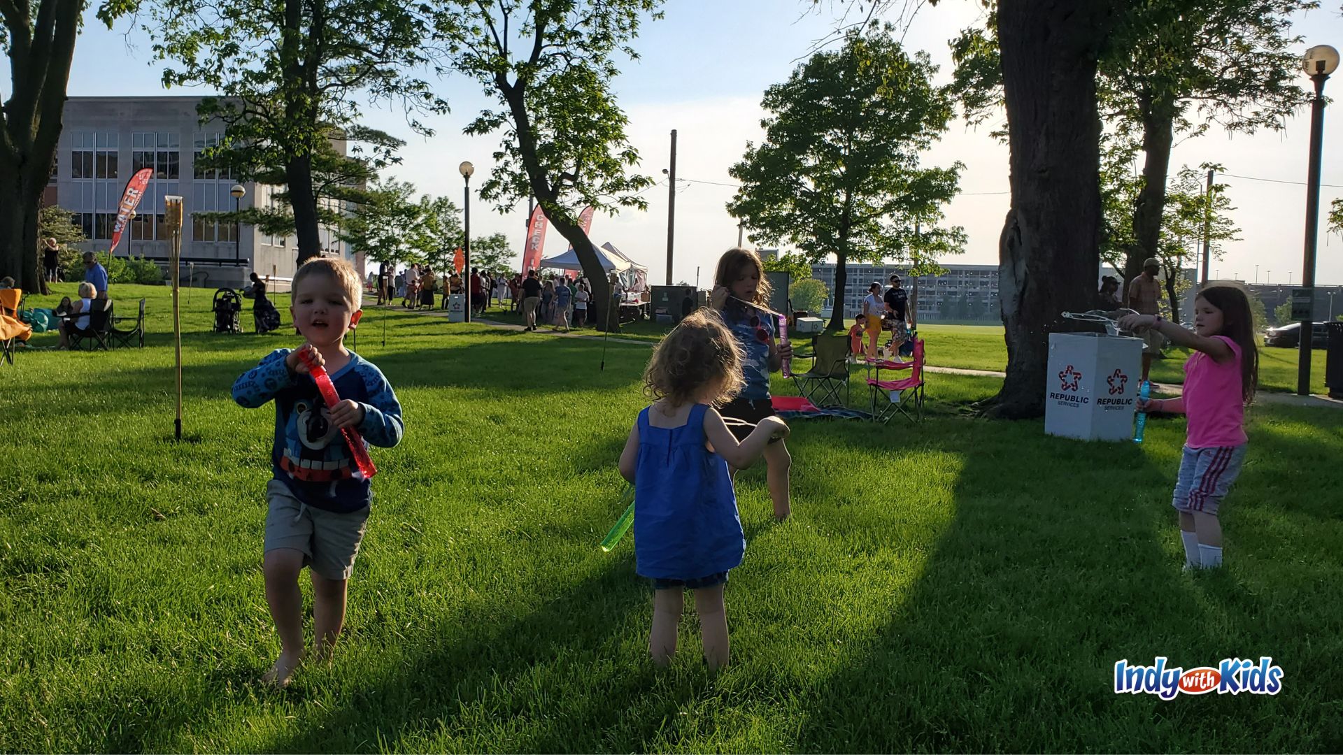 Young children play with glow sticks and bubbles on a grassy lawn during the Indianapolis Water Lantern Festival, with families, tents, and event activities behind them.