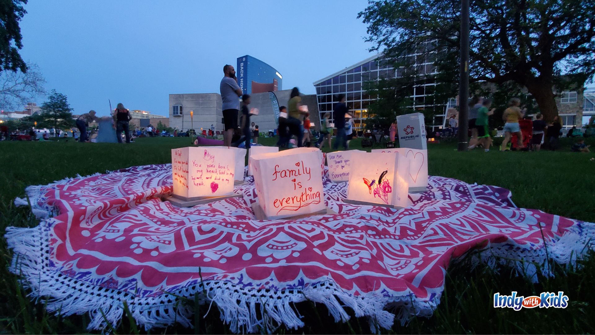 Glowing paper lanterns decorated with family messages rest on a pink picnic blanket in the grass at dusk, while festival guests gather near the water in the background.