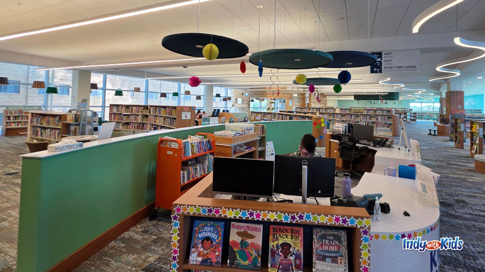 Wide view of the new Kid Central children’s section at Central Library, with a curved service desk, low bookshelves, colorful hanging decorations, large windows, and a sign pointing toward the Play and Story Time Lodge areas.