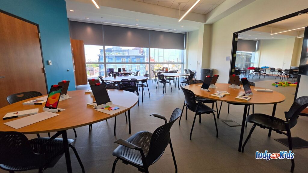 Bright classroom with large windows, round wooden tables, black chairs, and tablets arranged at each seat for children’s technology and STEAM programs.