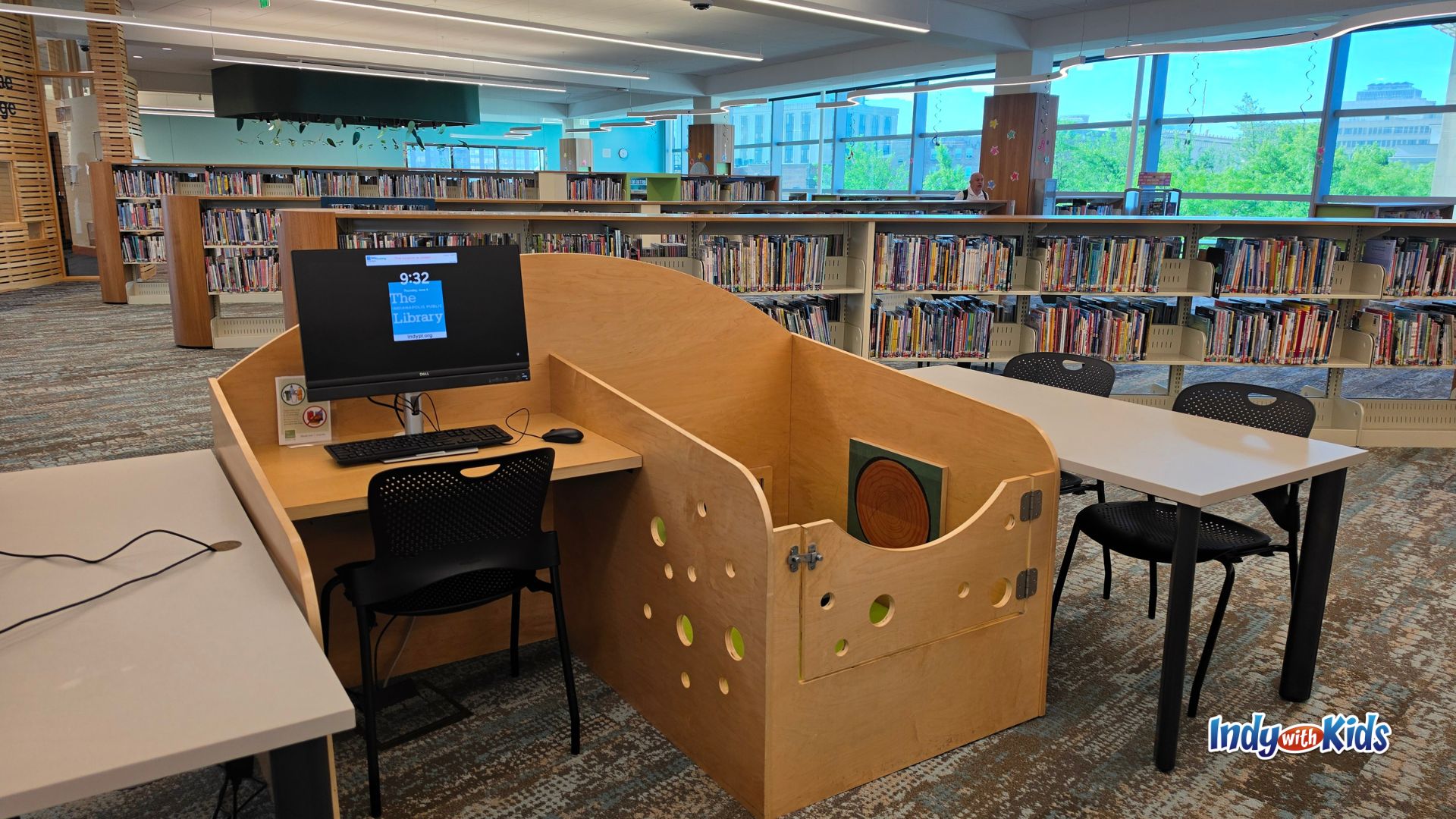 Wooden computer workstation surrounded by a small enclosed play area with circular cutouts, positioned beside tables and children’s bookshelves in Kid Central.