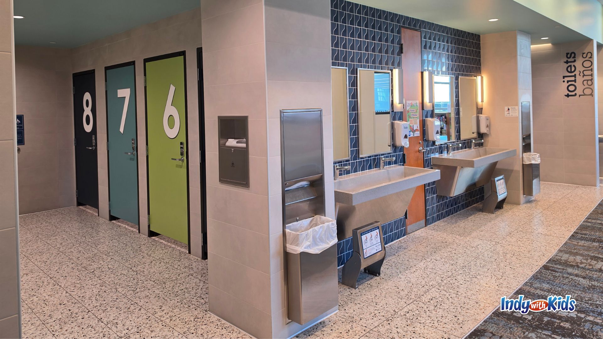 Family-friendly restroom area with numbered individual stall doors, two low open sinks, two standard-height sinks, mirrors, soap dispensers, a water fountain, and a bottle-filling station.