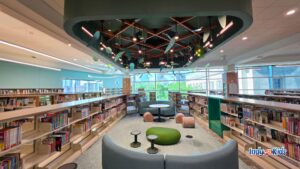 Cozy children’s reading area between low bookshelves, with curved seating, small tables, log-shaped stools, a green cushion, and decorative leaves hanging from a large ceiling canopy.