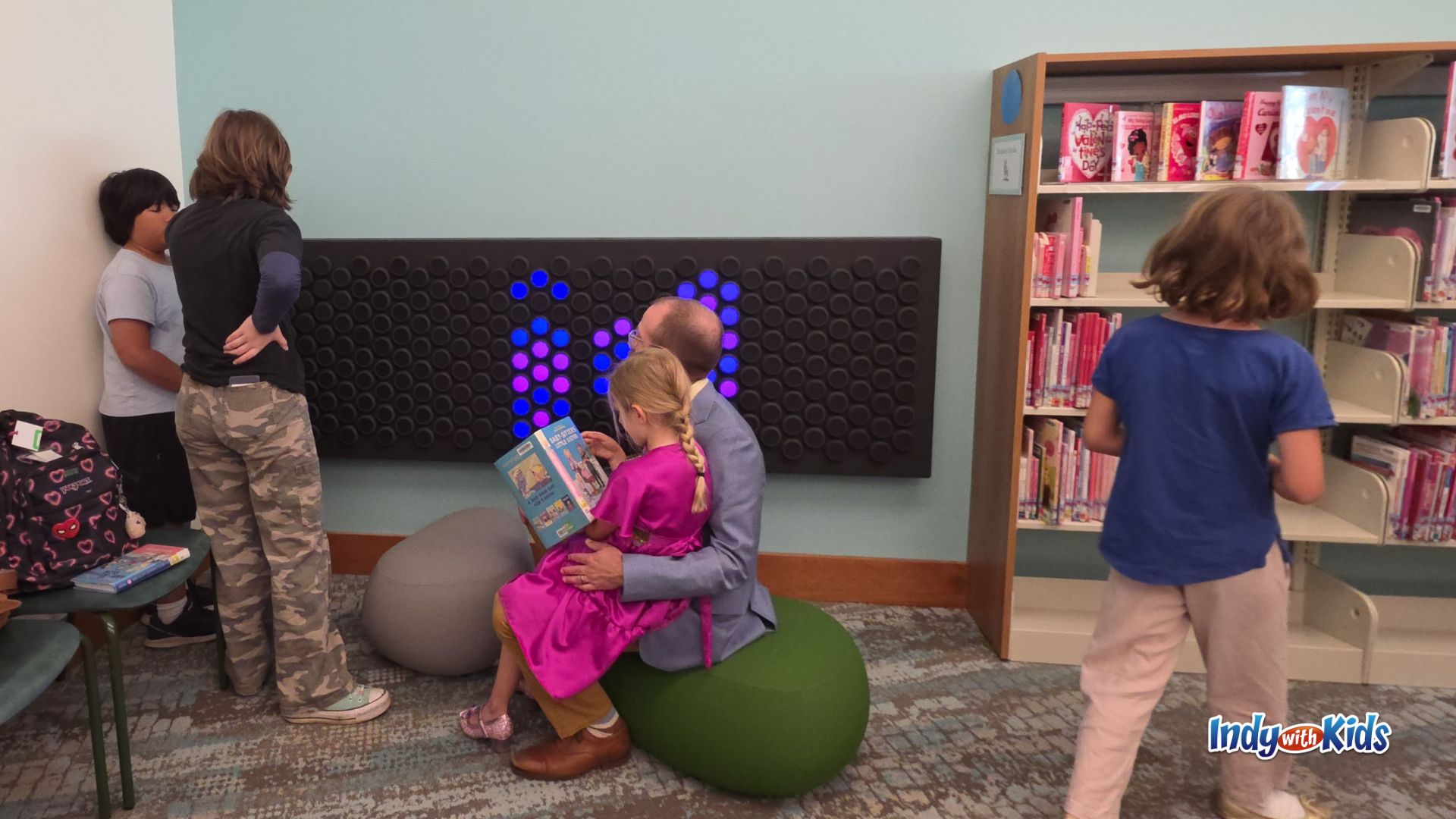 Children and an adult gathered around a large wall-mounted light board with glowing blue and purple pegs, while another child browses a nearby bookshelf.