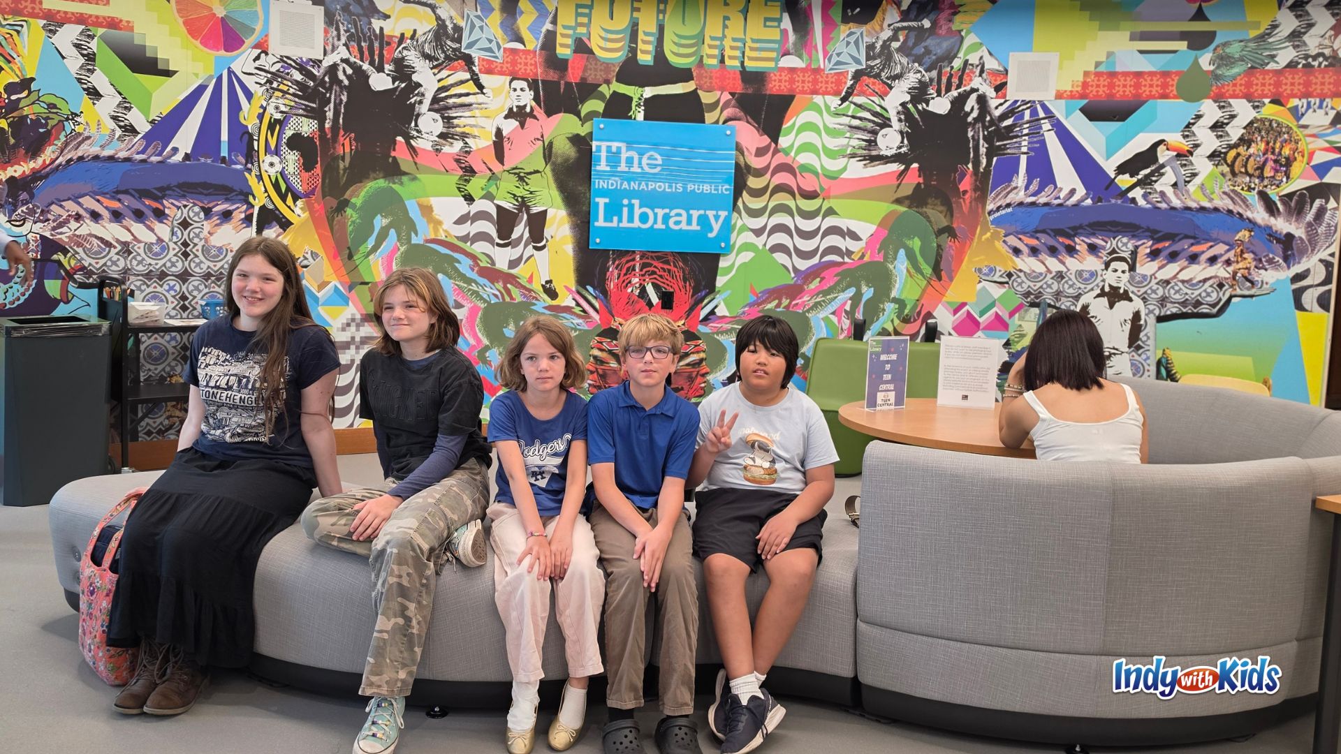 Five young people seated together on a curved gray couch in front of a colorful mural and an Indianapolis Public Library sign inside Teen Central.