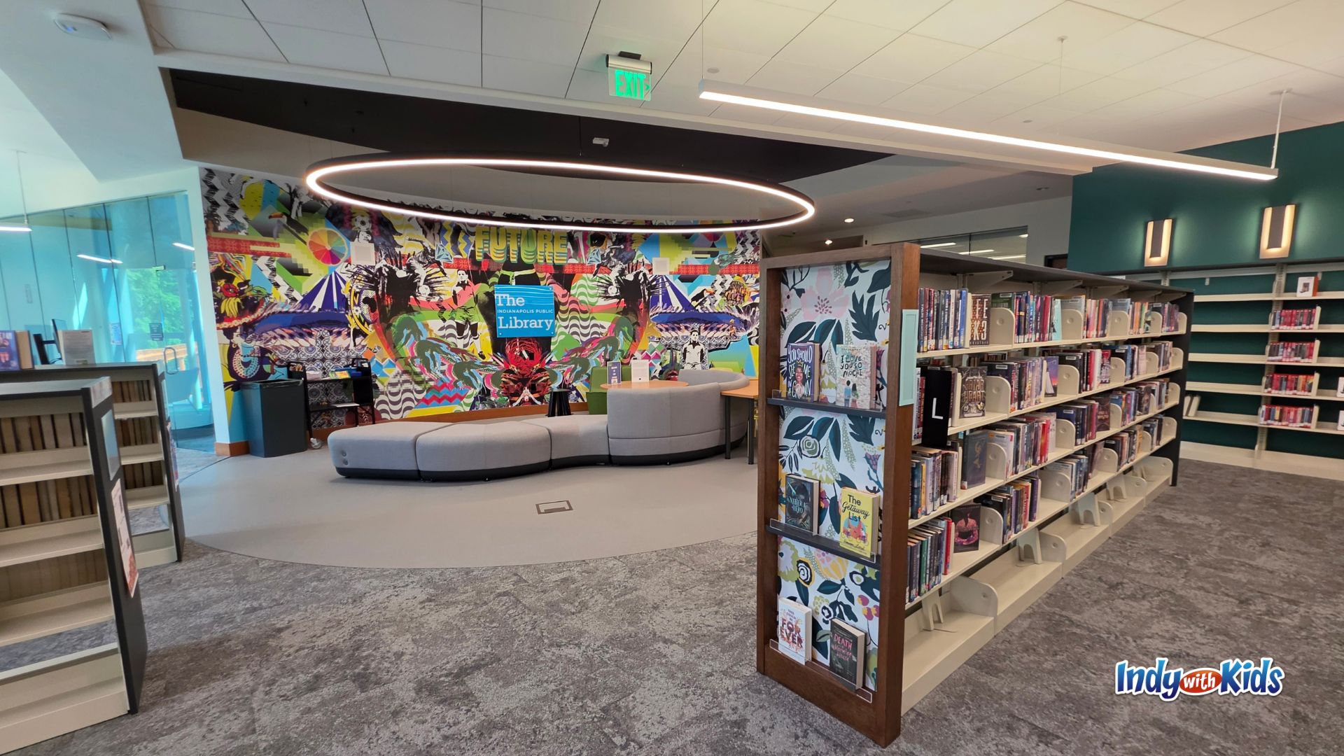 Wide view of the Teen Central reading area with bookshelves, a curved gray couch, a colorful mural, a large circular ceiling light, and teal accent walls.
