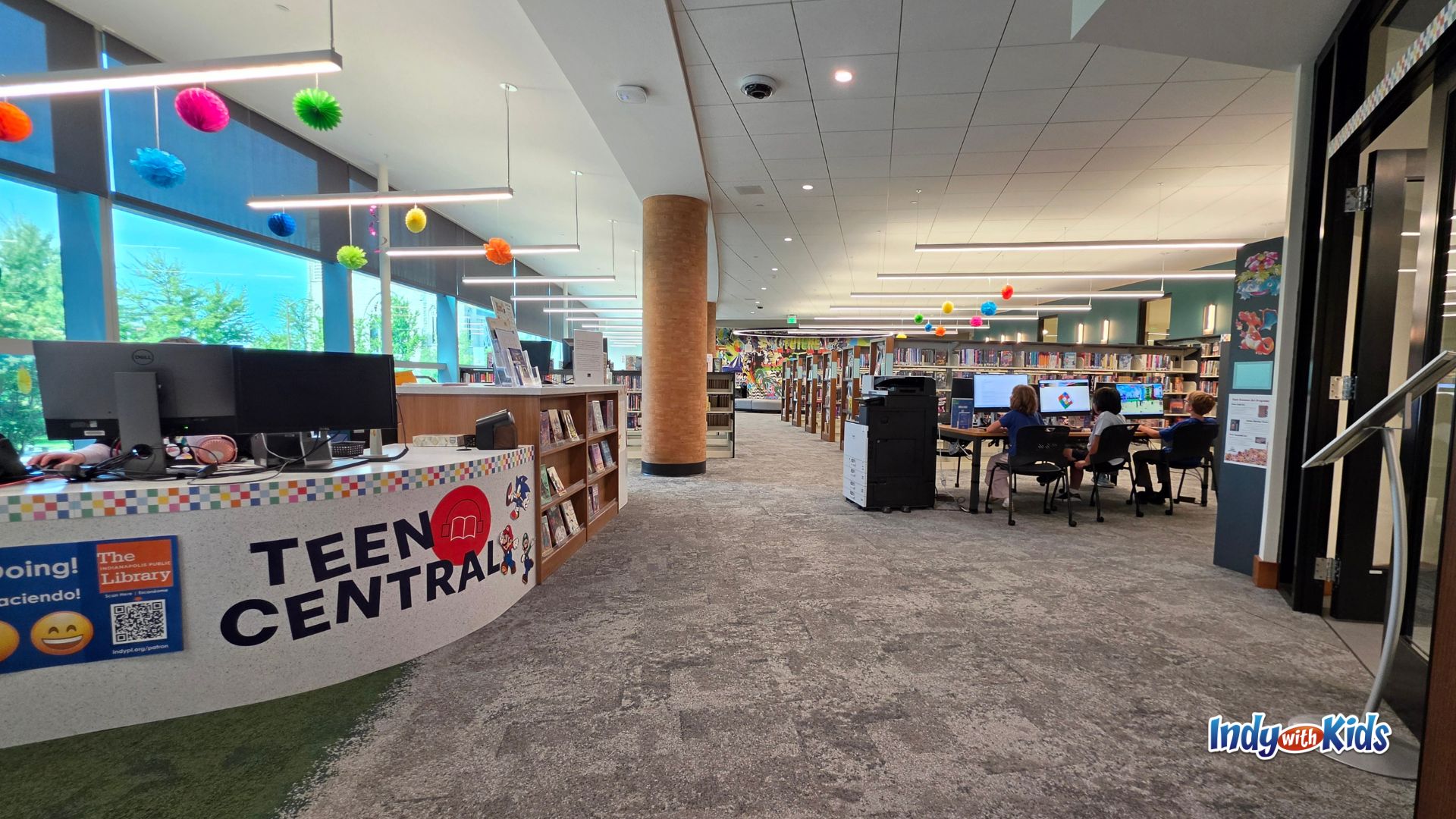 Wide view of the Teen Central entrance and service desk, with colorful hanging decorations, computer stations, bookshelves, large windows, and teens using computers in the distance.