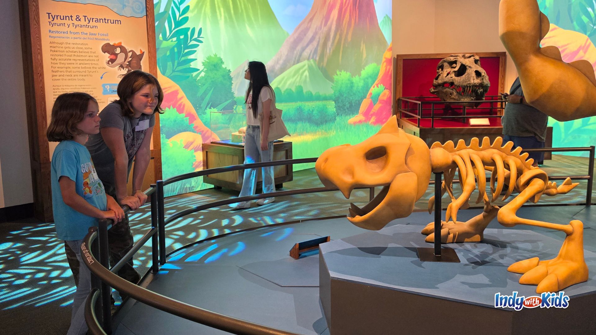 Two children look at a Tyrunt skeleton model inside the Pokémon Fossil Museum exhibit, with a T. rex skull and colorful prehistoric scenery in the background.