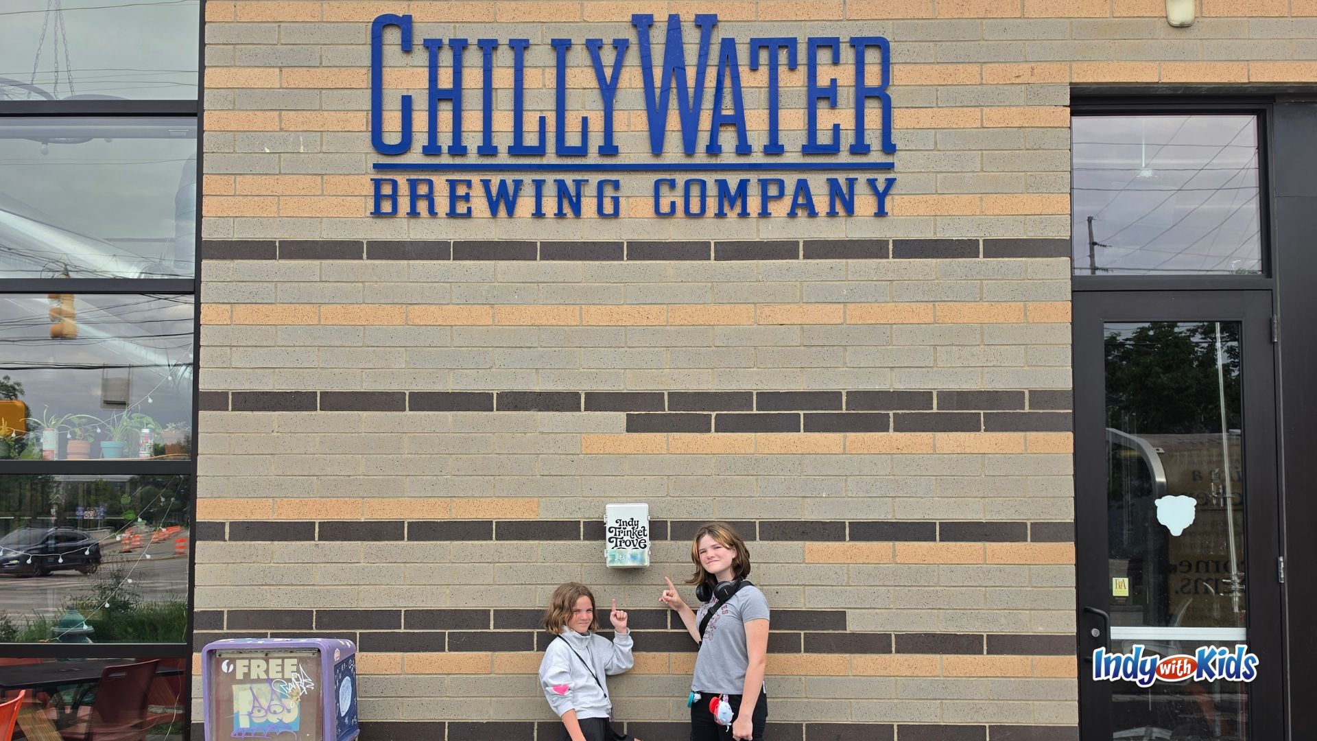 Two kids stand outside Chilly Water Brewing Company in Indianapolis, pointing to a small trinket trading box mounted on the brick wall.