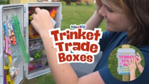 A child reaches into an open trinket trade box filled with small items, with large text over the image that reads “Trinket Trade Boxes.”