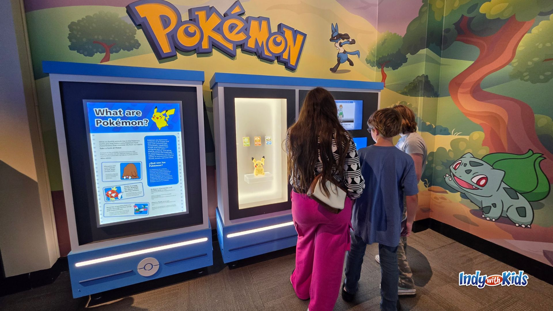 Children explore the entrance area of the Pokémon Fossil Museum exhibit, with colorful Pokémon graphics, informational displays, and a small Pikachu figure.