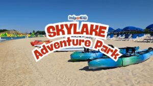 Text overlay reading “SKYLAKE Adventure Park” over a sunny beach scene with kayaks on white sand, blue umbrellas, lounge chairs, and water attractions in the distance.