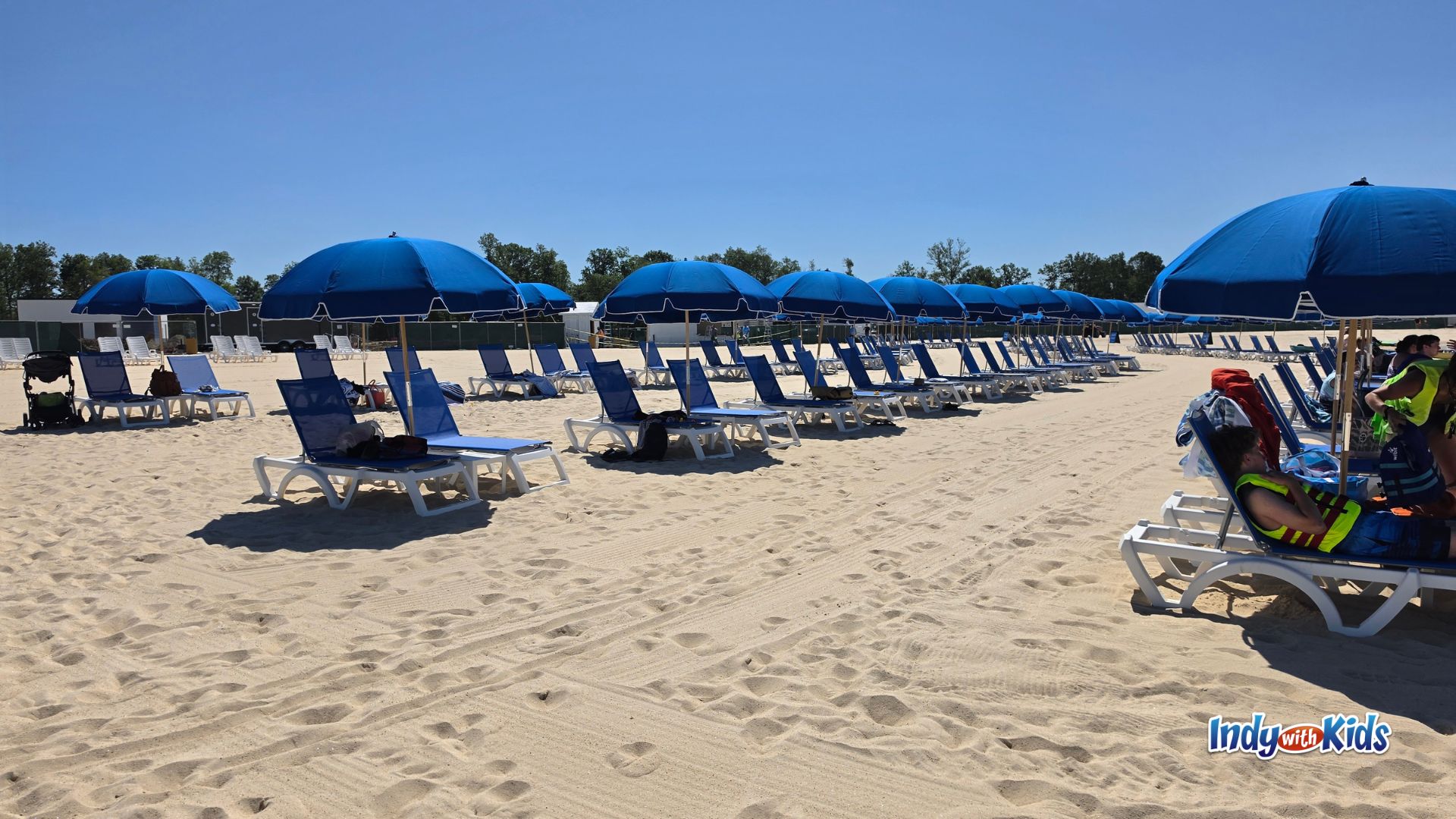 Rows of blue lounge chairs and matching blue umbrellas line the sandy beach at SKYLAKE Adventure Park on a bright sunny day.