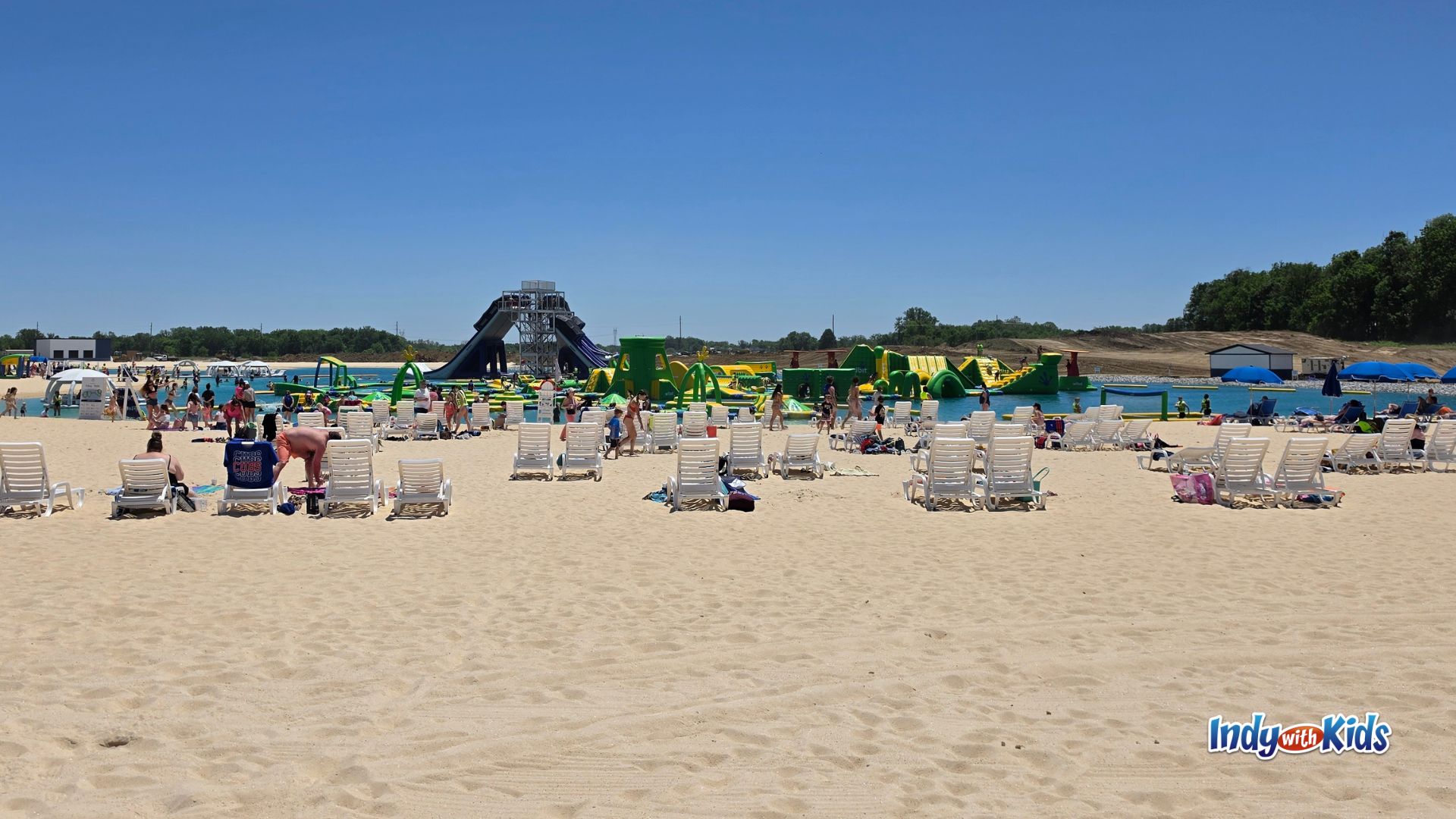 Wide view of the sandy beach and lake at SKYLAKE Adventure Park, with families relaxing in lounge chairs and kids playing on inflatable water obstacles in the background.